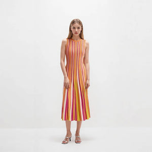 A woman stands against a plain white studio background wearing a sleeveless midi dress with vertical pink, orange, yellow, and white stripes, paired with metallic heeled sandals.