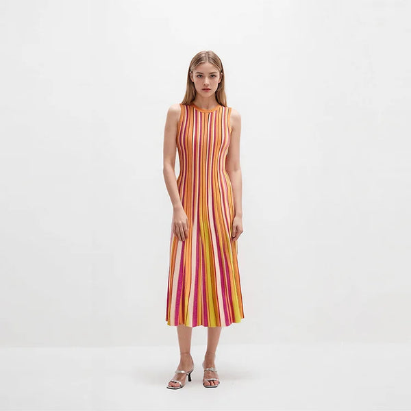 A woman stands against a plain white studio background wearing a sleeveless midi dress with vertical pink, orange, yellow, and white stripes, paired with metallic heeled sandals.
