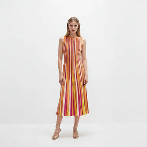 A woman stands against a plain white studio background wearing a sleeveless midi dress with vertical pink, orange, yellow, and white stripes, paired with metallic heeled sandals.