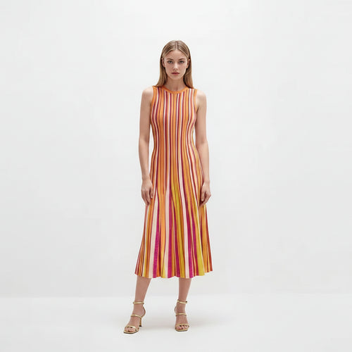 A woman stands against a plain white studio background wearing a sleeveless midi dress with vertical pink, orange, yellow, and white stripes, paired with metallic heeled sandals.