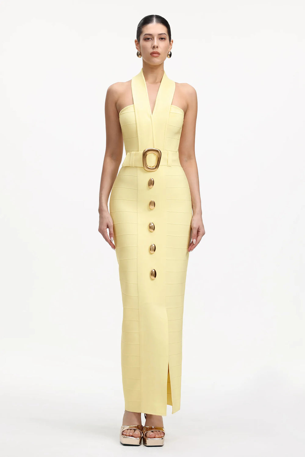 A model wears a pale yellow halter-neck midi dress with a structured bodice, a matching belt with a large gold buckle, and a front button detail, set against a plain white studio background.