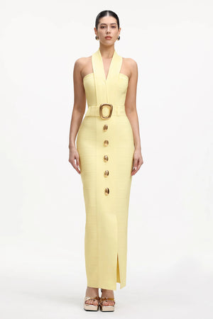 A model wears a pale yellow halter-neck midi dress with a structured bodice, a matching belt with a large gold buckle, and a front button detail, set against a plain white studio background.