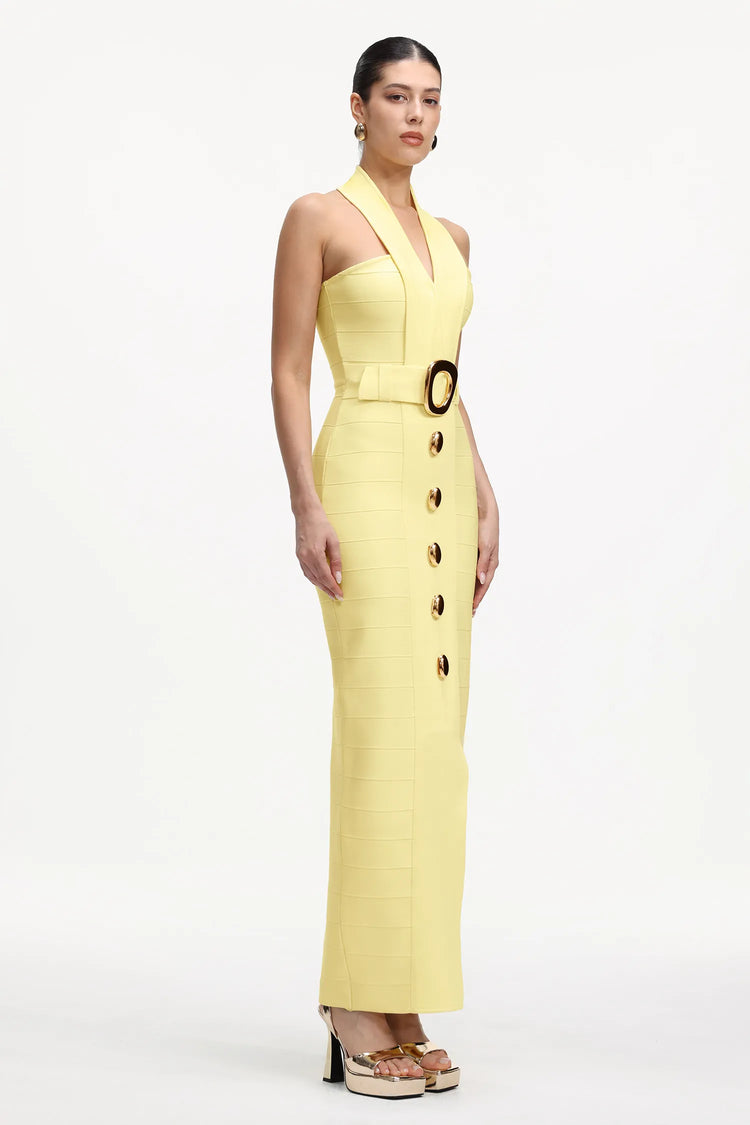 A model wears a pale yellow halter-neck midi dress with a structured bodice, a matching belt with a large gold buckle, and a front button detail, set against a plain white studio background.