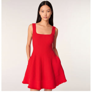 A model wearing a bright red ribbed knit sleeveless dress with a square neckline, wide shoulder straps, and a flared skirt with pockets, set against a plain light gray studio background.