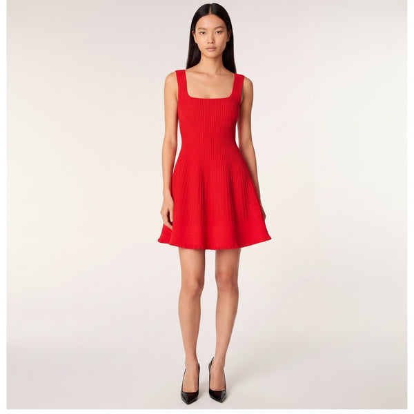 A model wearing a bright red sleeveless pleated dress with a square neckline and a flared skirt, set against a plain light gray studio background.