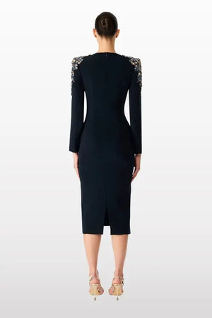 A woman viewed from the back wearing a fitted black knee-length dress with long sleeves and ornate embellished shoulder detailing, paired with beige high-heeled sandals against a light gray studio background.