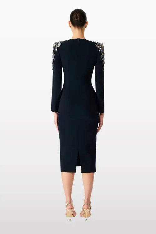 A woman viewed from the back wearing a fitted black knee-length dress with long sleeves and ornate embellished shoulder detailing, paired with beige high-heeled sandals against a light gray studio background.