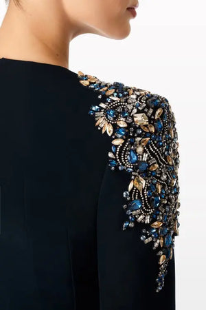 Close-up of a model wearing a black dress with elaborate shoulder embellishment featuring blue, gold, and clear rhinestones.
