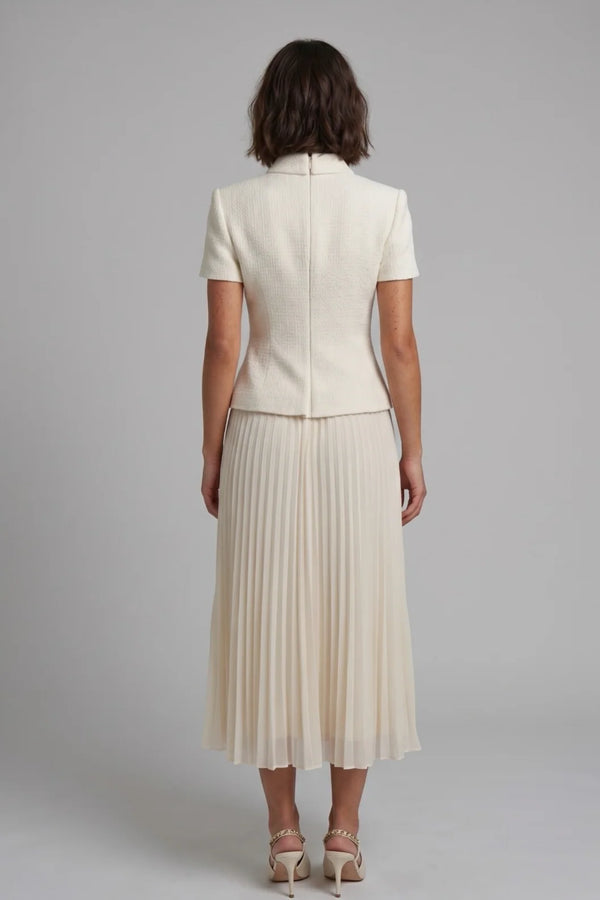 A woman viewed from the back wearing a cream-colored short-sleeve jacket and matching pleated midi skirt, paired with beige high-heeled sandals, against a solid gray studio background.