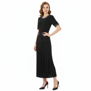 A woman modeling a black short-sleeve midi dress with silver rhinestone trim around the crew neck, sleeve cuffs, and waist, paired with a pleated skirt and black heels, on a solid white background.