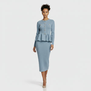 A model stands against a light gray studio background wearing a light blue ribbed knit two-piece set consisting of a long-sleeve cropped cardigan with a peplum hem and matching knee-length skirt.