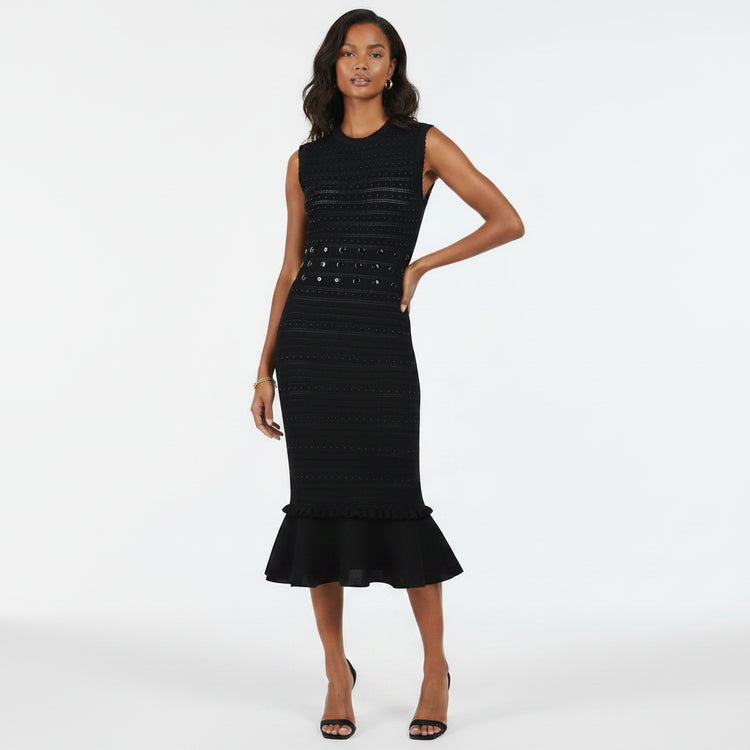 A woman modeling a sleeveless black midi dress with textured horizontal knit lines, a studded waistband, and a ruffled hem, set against a plain white studio background.