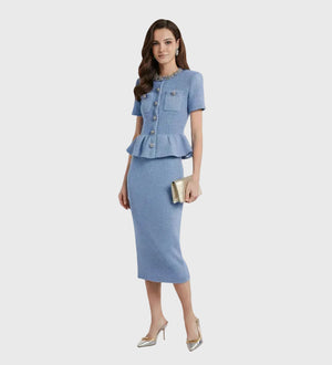 A woman models a light blue tweed two-piece set consisting of a short-sleeve peplum button-front jacket and a matching knee-length pencil skirt, styled with metallic pointed-toe heels and a small metallic clutch against a plain light gray studio background.