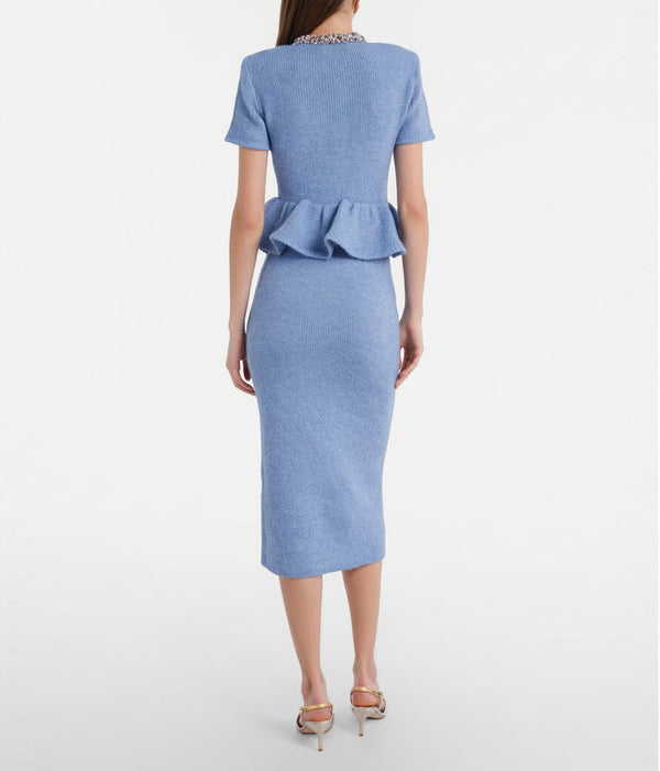 A woman viewed from the back wearing a light blue short-sleeve knit dress with a peplum waist and a beaded neckline, paired with metallic high-heeled sandals against a light gray studio background.