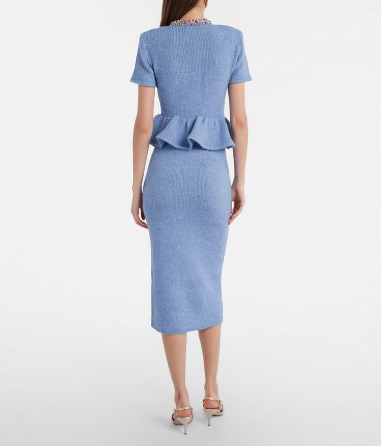 A woman models a light blue tweed two-piece set consisting of a short-sleeve peplum button-front jacket and a matching knee-length pencil skirt, styled with metallic pointed-toe heels and a small metallic clutch against a plain light gray studio background.