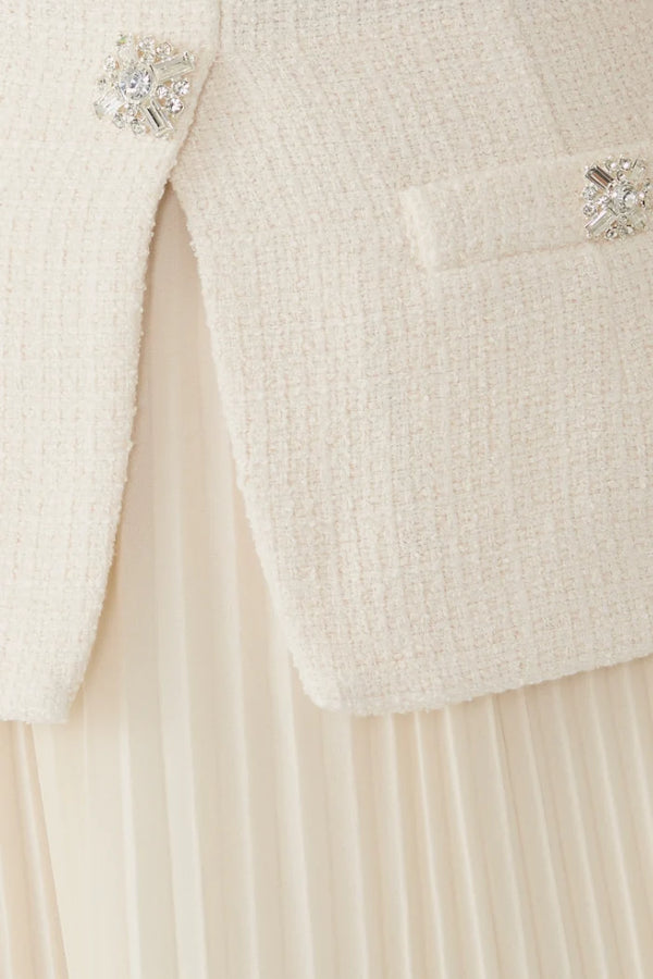 Close-up of a cream-colored tweed jacket with crystal-embellished buttons and a pleated cream skirt.