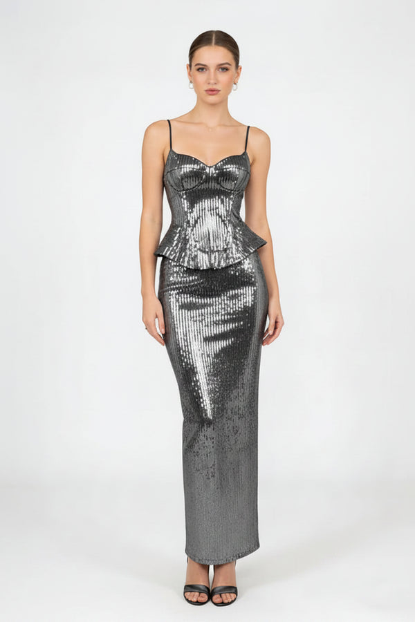 A woman modeling a floor-length, bodycon sequined dress in metallic silver with thin spaghetti straps and a sweetheart neckline, paired with a matching sequined clutch bag and black sandals, against a plain light gray studio background.