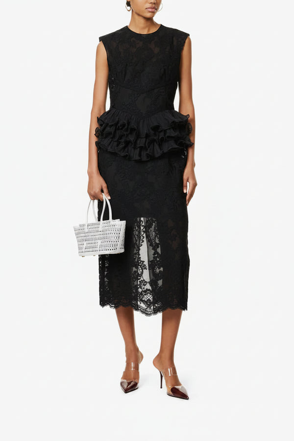 Woman wearing a black lace dress with ruffled details, holding a white woven handbag on a white background.