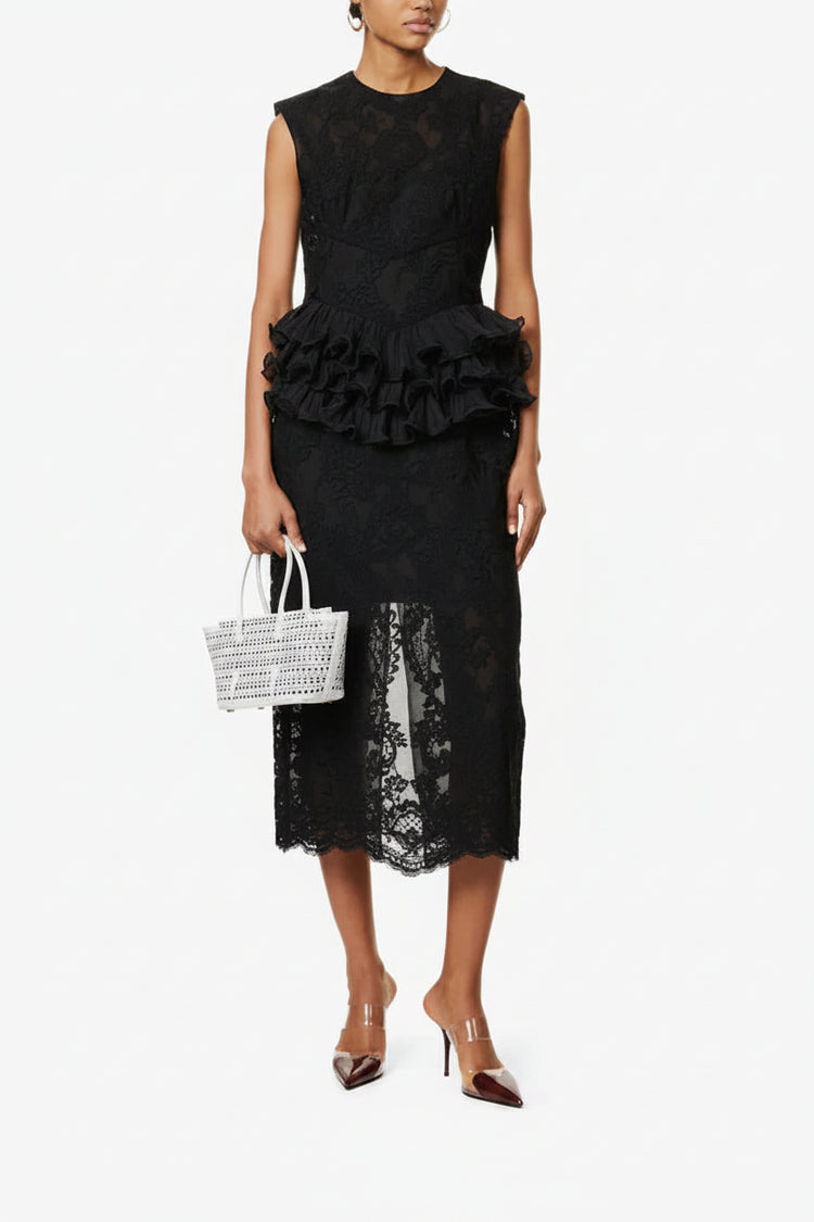Woman wearing a black lace dress with ruffled details, holding a white woven handbag on a white background.