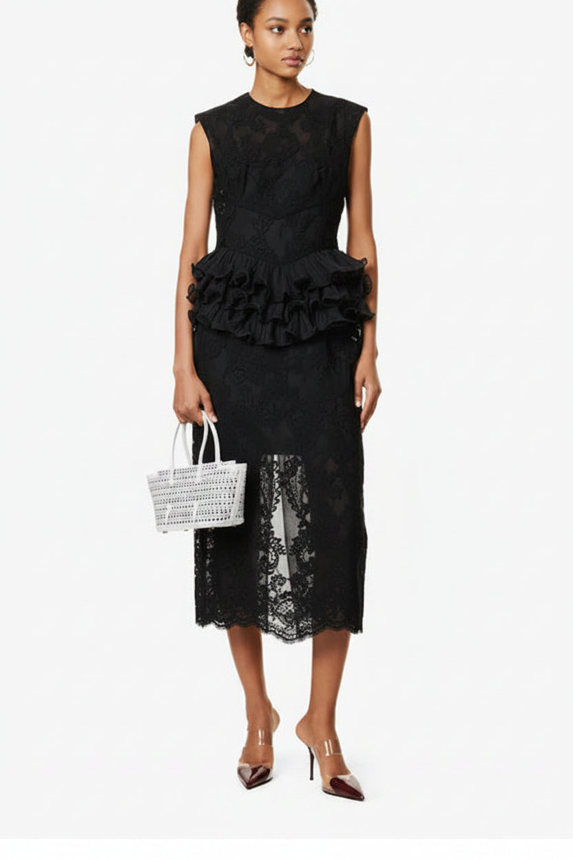 Woman wearing a black lace dress with ruffled details, holding a white woven handbag on a white background.