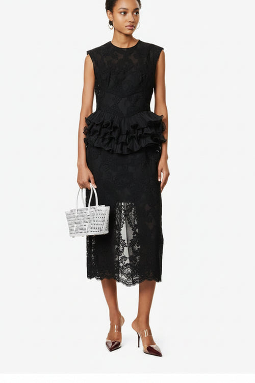 Woman wearing a black lace dress with ruffled details, holding a white woven handbag on a white background.
